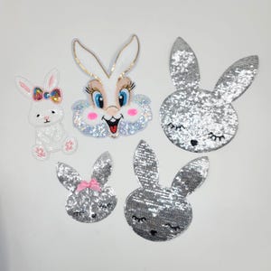 May include: A collection of five decorative bunny patches. One white bunny patch is embellished with sequins and a colorful bow. Three silver sequined bunny head patches and one bunny face patch with a pink bow are also included. The last patch is a bunny face with a smiling mouth.