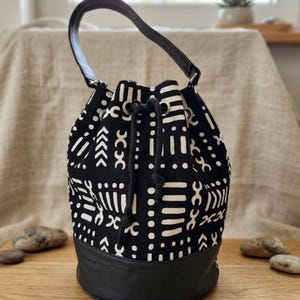 May include: A black and white patterned bucket bag with a black leather base and handle. The bag features a drawstring closure and a repeating geometric design. The bag is sitting on a wooden surface.
