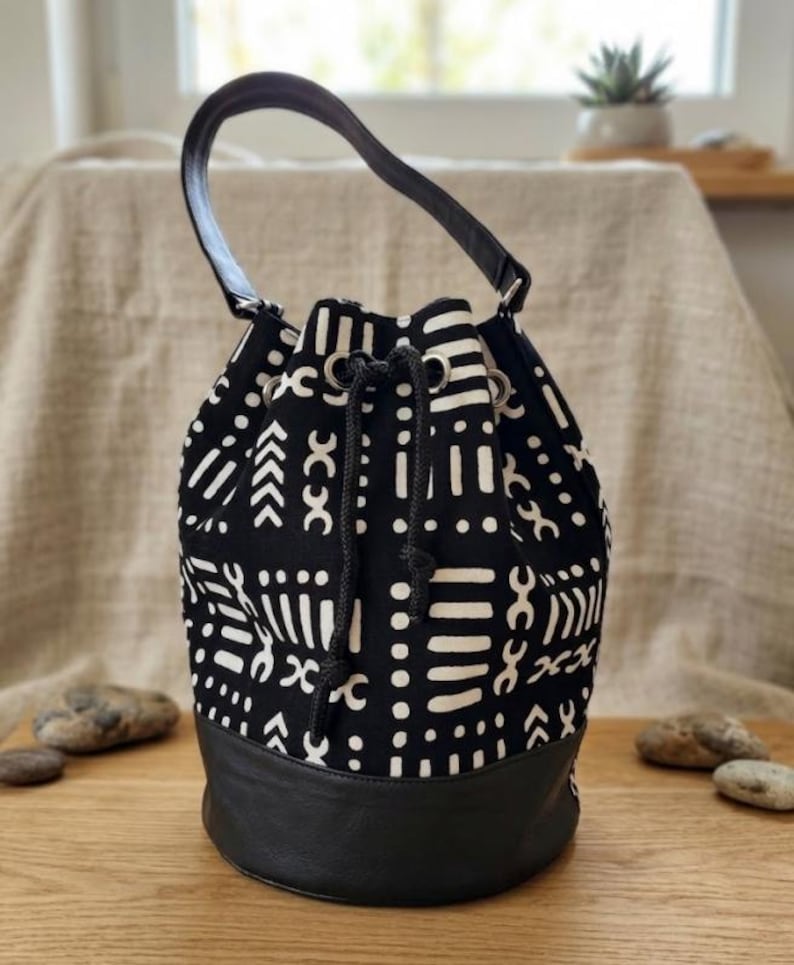 May include: A black and white patterned bucket bag with a black leather base and handle. The bag features a drawstring closure and a repeating geometric design. The bag is sitting on a wooden surface.