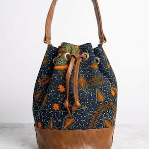 May include: A navy blue bucket bag with a brown leather base and handles. The bag's fabric features an orange and green floral pattern. The drawstring closure is secured with brown leather ties and gold-tone metal grommets.