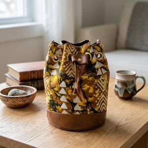 May include: A patterned bucket bag with a brown leather base and drawstring closure. The bag features a yellow, brown, and white geometric design. The bag is sitting on a wooden table.