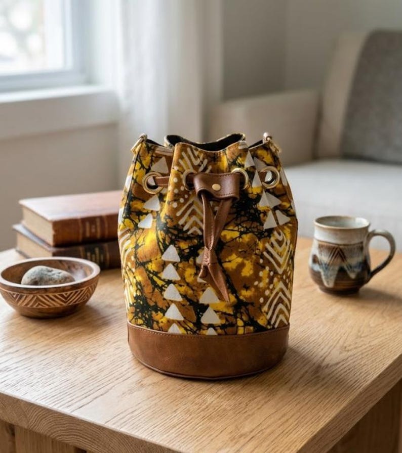 May include: A patterned bucket bag with a brown leather base and drawstring closure. The bag features a yellow, brown, and white geometric design. The bag is sitting on a wooden table.