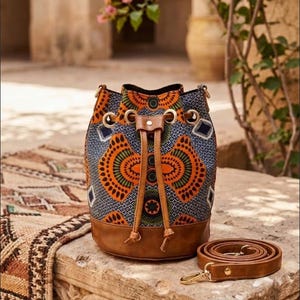 May include: A patterned bucket bag with a brown leather base and drawstring closure. The bag features a colorful geometric design in orange, green, and blue. A matching brown leather strap is coiled beside the bag.