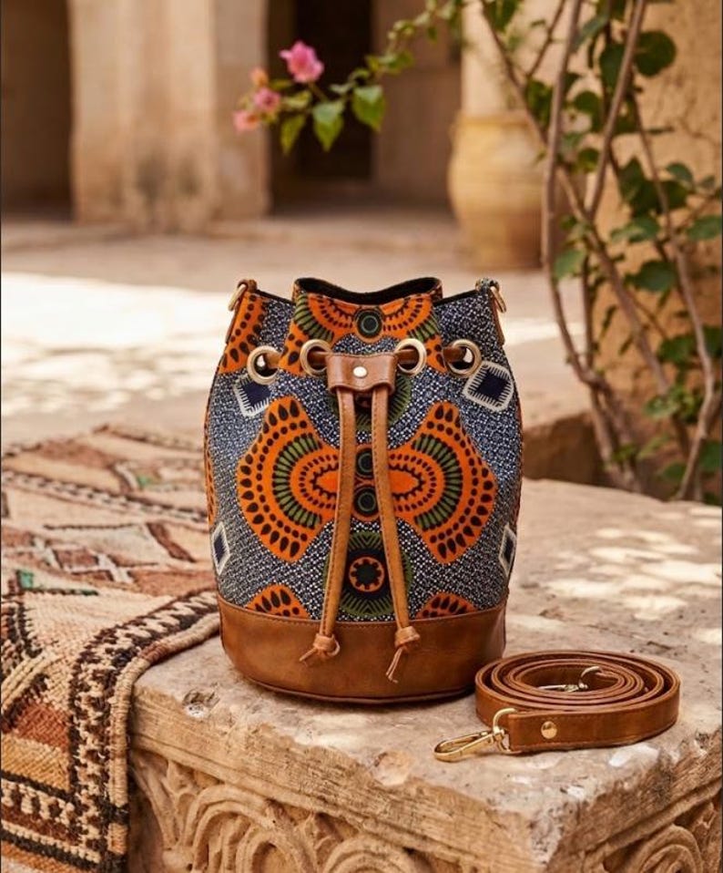 May include: A patterned bucket bag with a brown leather base and drawstring closure. The bag features a colorful geometric design in orange, green, and blue. A matching brown leather strap is coiled beside the bag.