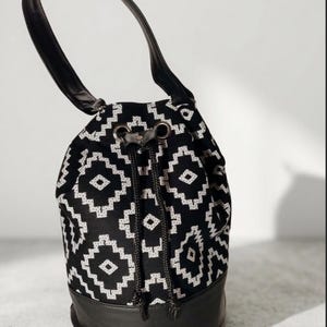 May include: Black and white patterned drawstring bag with a black leather base and handle. The bag features a geometric design and a drawstring closure. The handle is made of black leather.