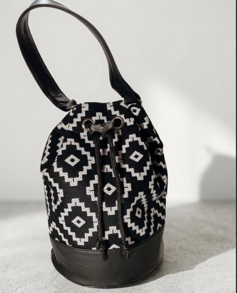 May include: Black and white patterned drawstring bag with a black leather base and handle. The bag features a geometric design and a drawstring closure. The handle is made of black leather.