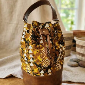 May include: A bucket bag with a brown leather base and handle. The bag's body features a yellow, black, and white patterned fabric. The drawstring closure and accents are brown leather. The bag is designed for carrying items.