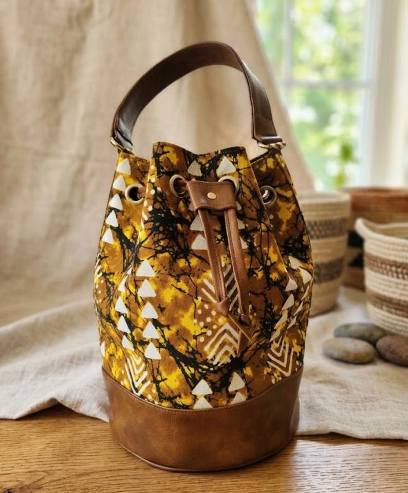 May include: A bucket bag with a brown leather base and handle. The bag's body features a yellow, black, and white patterned fabric. The drawstring closure and accents are brown leather. The bag is designed for carrying items.