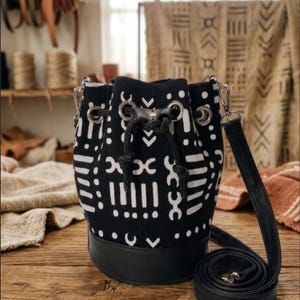 May include: Black and white patterned bucket bag with a black leather base and a long, adjustable strap. The bag features a drawstring closure and silver-tone metal hardware. The bag is sitting on a wooden surface.