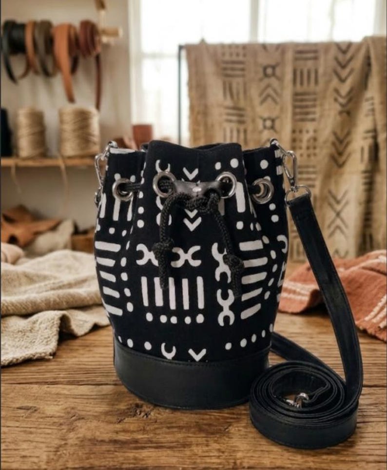 May include: Black and white patterned bucket bag with a black leather base and a long, adjustable strap. The bag features a drawstring closure and silver-tone metal hardware. The bag is sitting on a wooden surface.