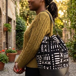 May include: A black and white patterned drawstring bag with a black leather bottom. The bag is carried over the shoulder. The bag's design features geometric shapes. The person is wearing a green sweater and black pants.