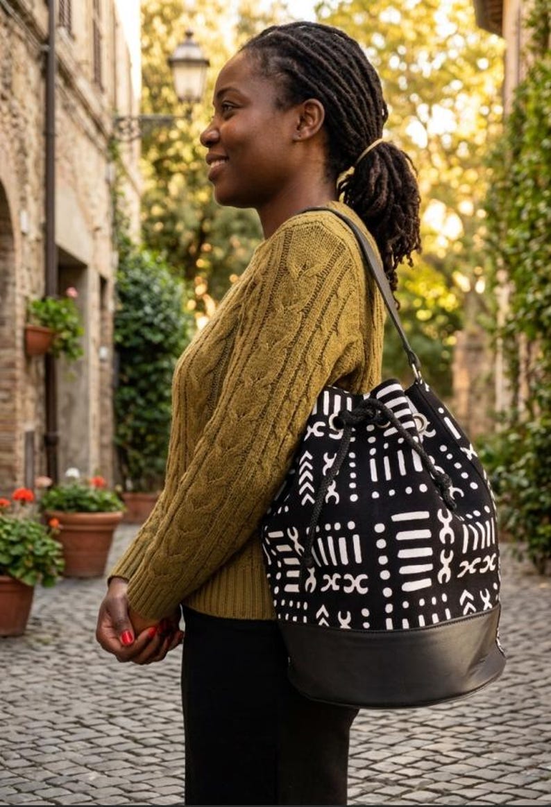 May include: A black and white patterned drawstring bag with a black leather bottom. The bag is carried over the shoulder. The bag's design features geometric shapes. The person is wearing a green sweater and black pants.