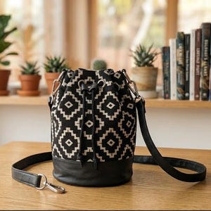 May include: A black and white geometric pattern bucket bag with a black leather base and strap. The bag has a drawstring closure and silver-tone hardware. The bag is sitting on a wooden table.