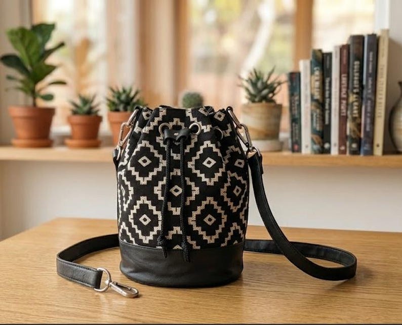 May include: A black and white geometric pattern bucket bag with a black leather base and strap. The bag has a drawstring closure and silver-tone hardware. The bag is sitting on a wooden table.