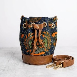May include: A navy blue bucket bag with an orange and green patterned design. The bag has a brown leather base, drawstring closure, and a detachable brown shoulder strap with gold-colored clasps.