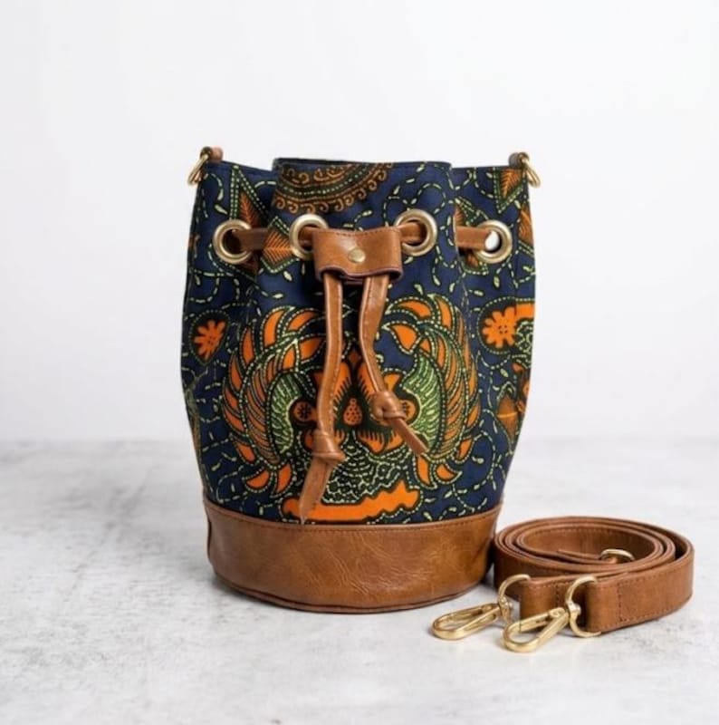May include: A navy blue bucket bag with an orange and green patterned design. The bag has a brown leather base, drawstring closure, and a detachable brown shoulder strap with gold-colored clasps.