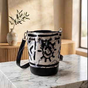 May include: A white and black bucket bag with a marble pattern and black accents. The bag has a black base, a drawstring closure, and a black shoulder strap. The bag is sitting on a marble surface.