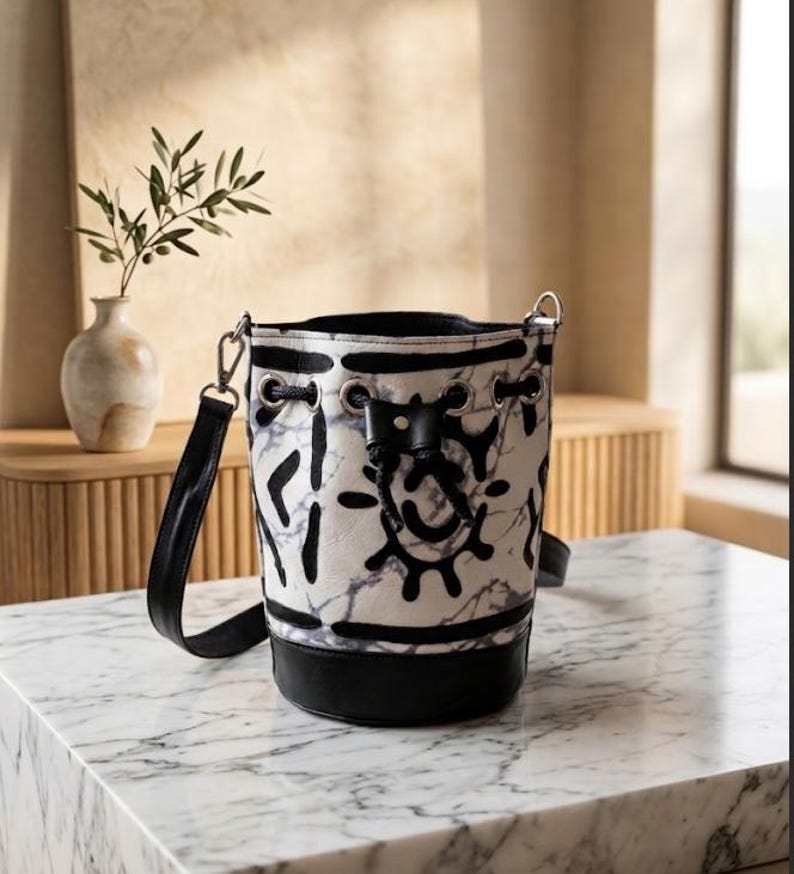 May include: A white and black bucket bag with a marble pattern and black accents. The bag has a black base, a drawstring closure, and a black shoulder strap. The bag is sitting on a marble surface.
