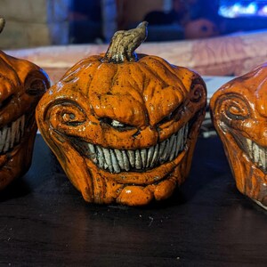 May include: Three orange ceramic pumpkin figurines arranged in a row. Each pumpkin features a carved, wide, toothy grin with visible white teeth, deep textured grooves, and a brown stem on top. The surface has a glossy finish with dark shading.