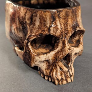 May include: A ceramic skull-shaped container with a wide opening. The skull is primarily beige with brown and dark brown accents, giving it a weathered appearance. The interior is dark brown. The container is likely used for decorative purposes.