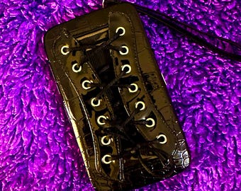 Könnte beinhalten: Schwarzes, rechteckiges Accessoire mit glänzendem, strukturiertem Finish, das einem Schuh ähnelt. Es hat goldfarbene Ösen und schwarze Schnürsenkel, mit einer schwarzen Kordel befestigt. Der Hintergrund ist ein leuchtend violettes, flauschiges Material.