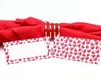Puede incluir: Dos tarjetas de lugar blancas con un borde de patrón de corazones rojos. Las tarjetas de lugar están sobre una servilleta roja con un anillo de servilleta dorado.