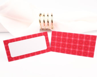 Puede incluir: Dos tarjetas de lugar rojas con un borde blanco y un fondo blanco. Las tarjetas de lugar tienen un patrón de pequeños corazones blancos sobre un fondo rojo. Una tarjeta de lugar tiene un espacio en blanco para escribir un nombre. Un anillo de servilleta dorado está en el fondo.