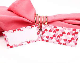 Puede incluir: Dos tarjetas de lugar rosas con un espacio en blanco para escribir un nombre. Las tarjetas de lugar tienen un patrón de corazones rosas y rojos. Una servilleta rosa está atada con un anillo de servilleta dorado.