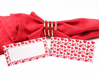 Puede incluir: Dos tarjetas de lugar con un patrón de corazones rojos. Las tarjetas de lugar están sobre una servilleta roja con un anillo de servilleta dorado.