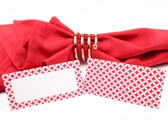 Puede incluir: Dos tarjetas de lugar rojas con diseños geométricos blancos. Las tarjetas de lugar están en blanco y listas para escribir. Una servilleta roja con un anillo de servilleta dorado está en el fondo.