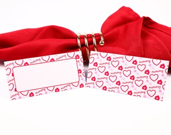 Puede incluir: Dos tarjetas de lugar con un fondo rosa y blanco con un patrón repetido de corazones rojos y la palabra "mamá". Las tarjetas de lugar están sobre una servilleta roja con un anillo de servilleta dorado.