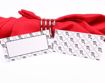 Puede incluir: Dos tarjetas de lugar blancas con un patrón blanco y negro de la letra "M" y un borde negro. Una tarjeta tiene un espacio en blanco para escribir un nombre. Las tarjetas están sobre una servilleta roja con un anillo de servilleta plateado.