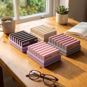 May include: Four rectangular desk organizers with vertical slots, each featuring the text MON and a small butterfly icon. The organizers are colored in dark gray, beige, light pink, and lavender, with each base matching the color of its slots.