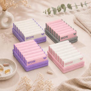 Könnte beinhalten: Vier pastellfarbene Pillenboxen werden gezeigt, jede mit einem anderen Farbschema. Die Boxen sind mit "MON" beschriftet und haben ein Schmetterlingsdesign. Die Boxen sind zur Aufbewahrung von Pillen und Vitaminen gedacht.