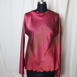 Puede incluir: Una blusa de manga larga de color burdeos con cuello y dobladillo con volantes. La blusa está hecha de un material sedoso y tiene un sutil efecto degradado.