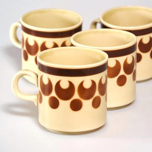 May include: Four cream-colored ceramic mugs with brown crescent and dot patterns. Each mug has a brown band around the top and a curved handle. The mugs are arranged in a group, showcasing their retro design.