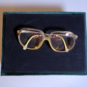 Può includere: Un paio di occhiali vintage con montatura marrone chiaro e lenti trasparenti. Gli occhiali sono appoggiati su una superficie di velluto verde scuro e nero. Le montature hanno una forma rettangolare classica.