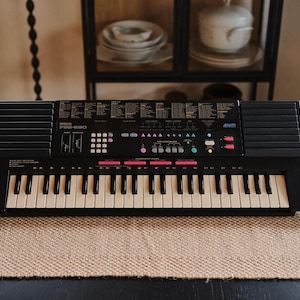 Puede incluir: Un teclado Yamaha PSR-580 negro con teclas blancas y negras. El teclado tiene varios botones y una pantalla. Se coloca sobre una estera tejida de color beige.