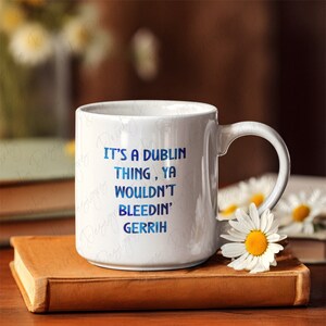 Könnte beinhalten: Weiße Keramiktasse mit dem Text "It's a Dublin Thing, Ya Wouldn't Bleedin' Gerrih" in blauem Farbverlauf. Die Tasse steht auf einem braunen Buch, mit Gänseblümchen und anderen Büchern im Hintergrund.