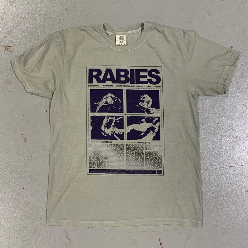 May include: A light green t-shirt with a graphic print of a newspaper article about rabies. The article includes the words "Rabies", "Incubation", "Prodrome", "Acute Neurological Period", "Coma", "Death", "Furious", and "Paralytic".