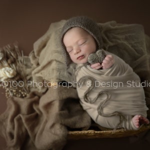 May include: A newborn baby is swaddled in a beige blanket and wearing a brown knitted hat. The baby is holding a small gray heart-shaped toy. The baby is lying in a woven basket on a brown background.
