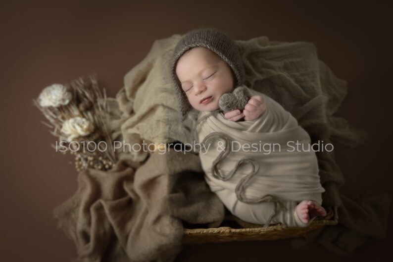 May include: A newborn baby is swaddled in a beige blanket and wearing a brown knitted hat. The baby is holding a small gray heart-shaped toy. The baby is lying in a woven basket on a brown background.
