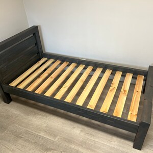 以下が含まれることがあります： ヘッドボードとフットボードを備えた、濃い灰色の木製ベッドフレーム。 ベッドフレームは、天然木で作られたスラットベースを備えています。 ヘッドボードは角度がついており、水平スラットがあります。 ベッドフレームはシングルサイズのマットレス用に設計されています。