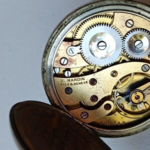 May include: An open antique pocket watch revealing its internal mechanisms. The gold-toned face displays silver gears and the inscription "U. NARDIN LOCLE & GENEVE". The silver-coloured watch case is partially visible.