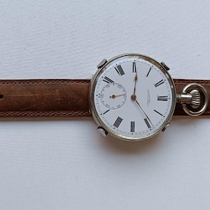 May include: A vintage pocket watch with a white face, black Roman numerals, and a small seconds dial. The watch is attached to a brown leather strap with a silver buckle. The watch face is approximately 5 cm in diameter.