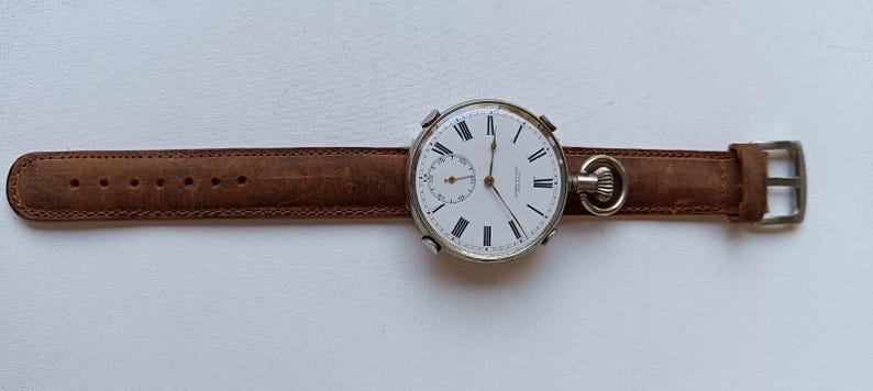 May include: A vintage pocket watch with a white face, black Roman numerals, and a small seconds dial. The watch is attached to a brown leather strap with a silver buckle. The watch face is approximately 5 cm in diameter.