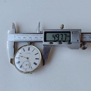 May include: A vintage pocket watch with a white face, black Roman numerals, and a subdial. The watch is being measured with a digital caliper, showing a measurement of 4.977 mm.