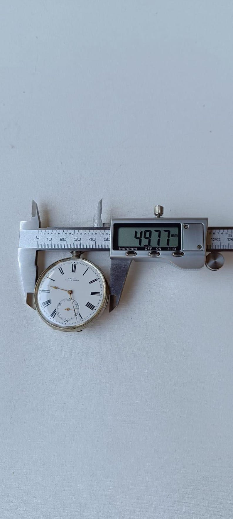May include: A vintage pocket watch with a white face, black Roman numerals, and a subdial. The watch is being measured with a digital caliper, showing a measurement of 4.977 mm.