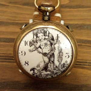 Op de afbeelding: Antieke zakhorloge met een goudkleurige kast en een witte wijzerplaat met een zwart-wit illustratie van een cherubijn. Het horloge heeft een bruine leren band en een goudkleurige opwindkroon. De achtergrond is van hout.