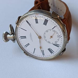 May include: A vintage pocket watch with a white face, black Roman numerals, and a small seconds dial. The watch has a silver-coloured case and a brown leather strap. The watch measures approximately 5 cm in diameter.